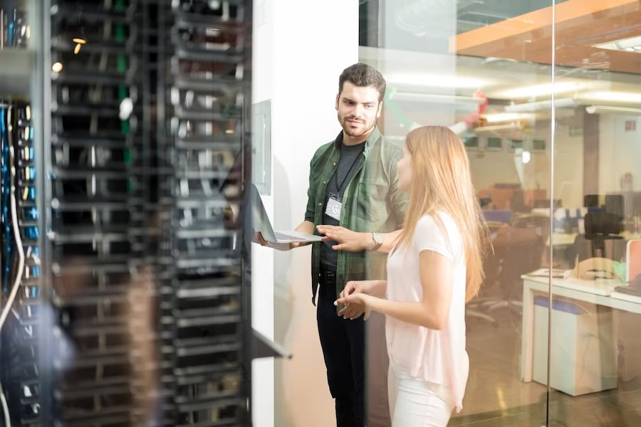 A man with a laptop in front of a server, accompanied by a woman